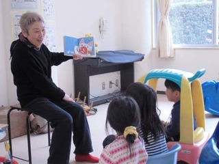遊具やイスに座った子ども達の前で絵本の読み聞かせをする女性の様子