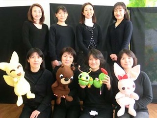 手作りの人形と並ぶ劇団員の女性8人の写真